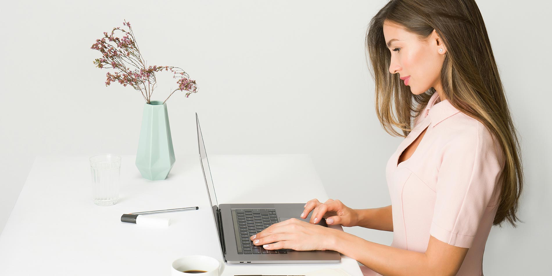 Profile view of a woman with long hair typing on a laptop at a clean white desk